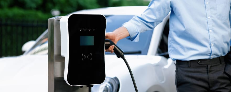 Closeup progressive suit-clad businessman with his electric vehicle recharge his car on public charging station in modern city with power cable plug and renewable energy-powered electric vehicle.