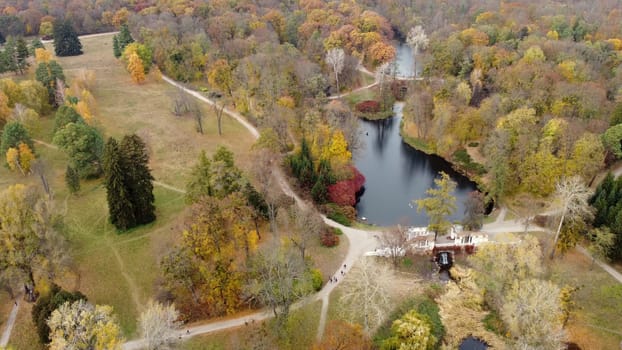 Beautiful landscape view of autumn park with lakes with swans, trees with yellow red green leaves, meadow, architecture, waterfall and people walking along dirt paths on autumn day. Natural background