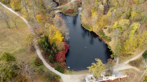 Beautiful landscape view of autumn park with trees with yellow fallen leaves, lakes, architecture and people walking along dirt paths on autumn day. Flying over autumn park scenery. Top view.