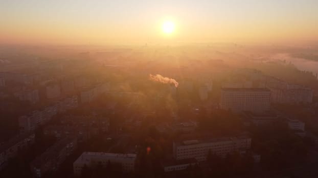 Beautiful cityscape in summer at sunrise. Flying over many multi-storey buildings and a chimney with white smoke. Bright shining sun. Urban landscape. Dawn sunrise dawning sunset sundown, rising sun.