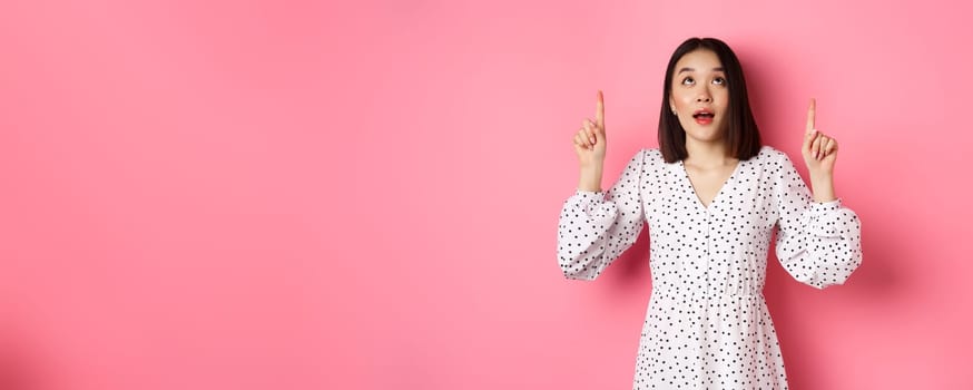 Image of beautiful asian girl in dress looking, pointing fingers up, gazing dreamy at promo offer, standing over pink background.