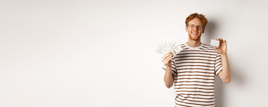 Shopping and finance concept. Young redhead man with beard and glasses showing plastic credit card with money in dollars, white background.