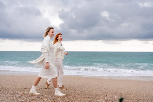 Women sea walk friendship spring. Two girlfriends, redhead and blonde, middle-aged walk along the sandy beach of the sea, dressed in white clothes. Against the backdrop of a cloudy sky and the winter sea. Weekend concept