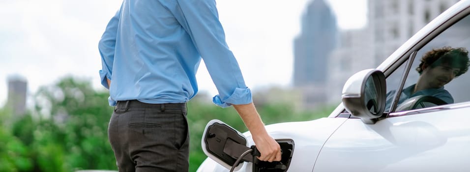 Closeup progressive man holding EV charger plug from public charging station for electric vehicle with background of residential building as concept eco-friendly sustainability energy car concept.