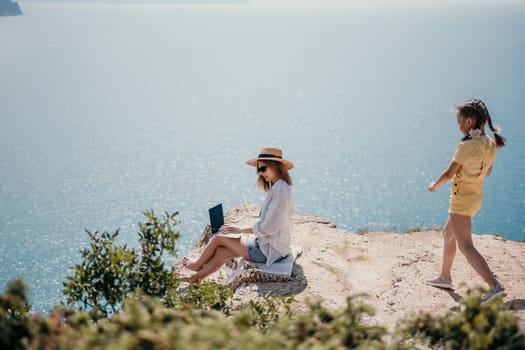 Successful business woman in yellow hat working on laptop by the sea. Pretty lady typing on computer at summer day outdoors. Freelance, travel and holidays concept.