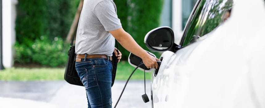 Progressive asian man install cable plug to his electric car with home charging station in the backyard. Concept use of electric vehicles in a progressive lifestyle contributes to clean environment.