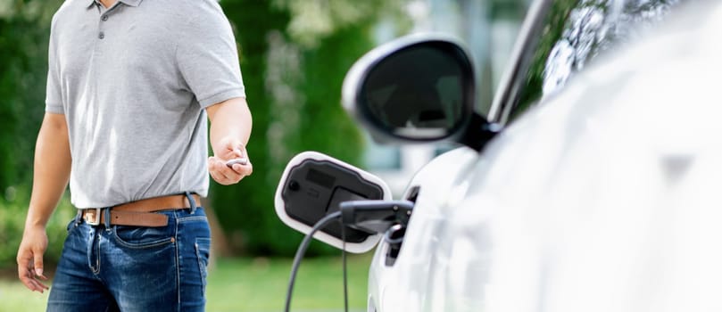 Progressive asian man install cable plug to his electric car with home charging station in the backyard. Concept use of electric vehicles in a progressive lifestyle contributes to clean environment.