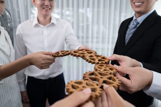 Closeup hand holding wooden gear by businesspeople wearing suit for harmony synergy in office workplace concept. Group of people hand making chain of gears into collective form for unity symbol.