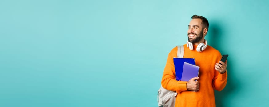 Education. Handsome male student with headphones and backpack, using mobile phone and holding notebooks, smiling happy, standing over turquoise background.