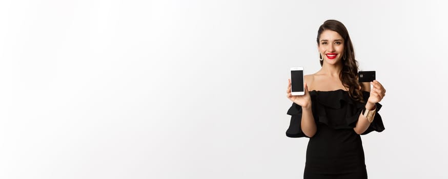 Online shopping concept. Fashionable brunette woman in black dress, showing mobile screen and credit card, smiling pleased, standing over white background.