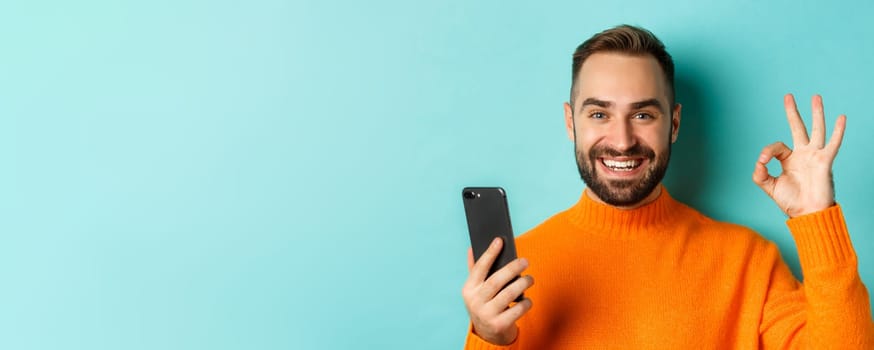 Close-up of bearded man holding smartphone and showing okay sign, recommending mobile app, standing satisfied over turquoise background.