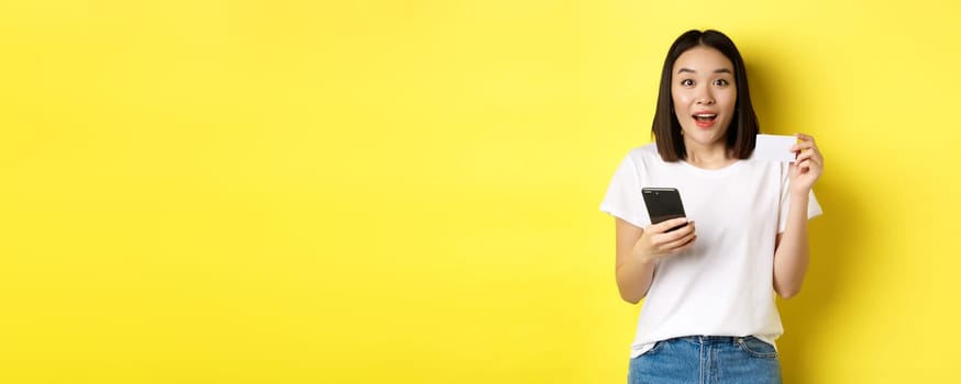 E-commerce and online shopping concept. Excited asian woman order in internet, holding smartphone and plastic credit card, standing over yellow background.