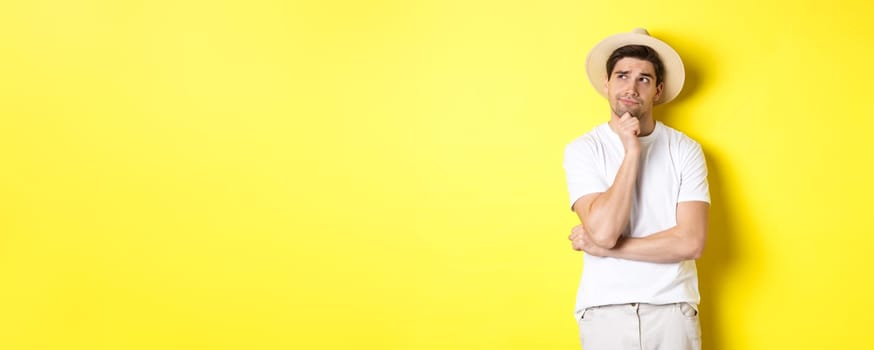 Concept of tourism and summer. Thoughtful man tourist pondering, looking at upper left corner and thinking, standing in straw hat and white t-shirt against yellow background.
