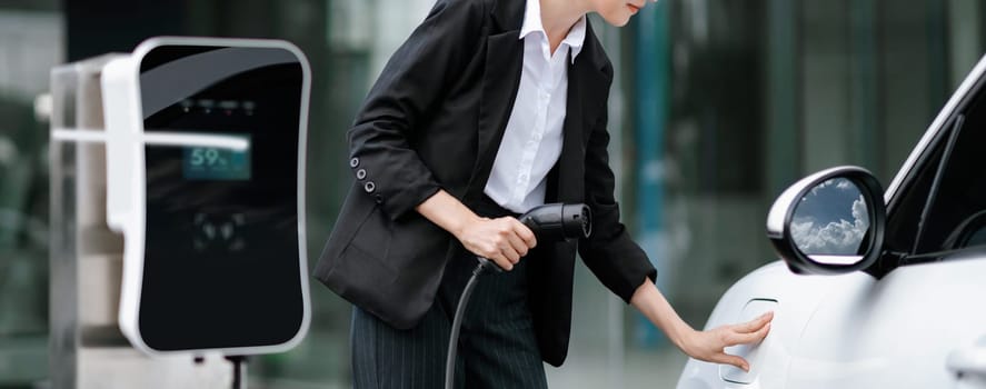 Progressive businesswoman insert charger plug from charging station to her electric vehicle with apartment condo building in background. Eco friendly rechargeable car powered by sustainable energy.