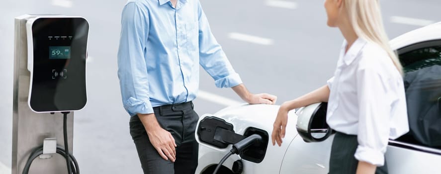 Progressive businessman and businesswoman with electric car parking and connected to public charging station before driving around city center. Eco friendly rechargeable car powered by clean energy.