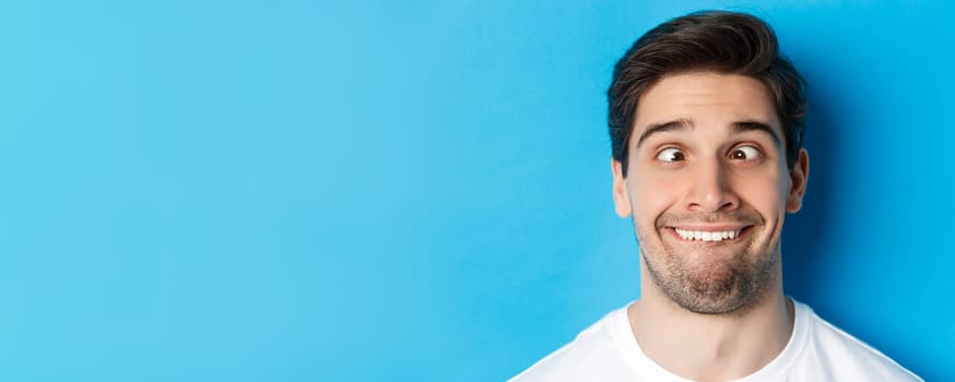 Head shot of young man making funny expressions, smiling and squinting, standing over blue background.