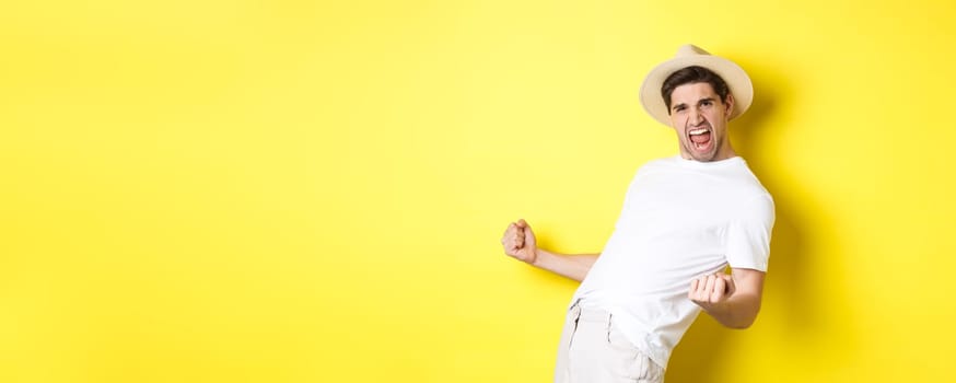 Concept of tourism and vacation. Satisfied young male tourist celebrating, winning something and rejoicing, making fist pump and shouting yes, standing against yellow background.