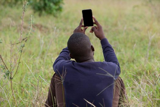 young man sitting back on the grass in the park holds in the air and looks on phone in hand