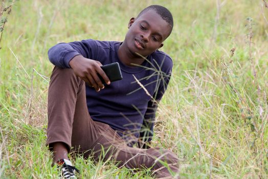 young smiling man sitting on the grass in the park is using and watching on mobile phone
