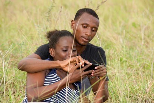 Young african couple sitting in the grass looking at the mobile phone together.