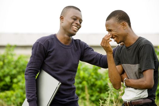 two young student chatting and laughing standing outdoors with a computer in their hands