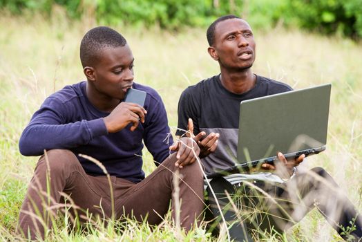 two happy young men talk, work in front of a laptop and relax while sitting in the park.
