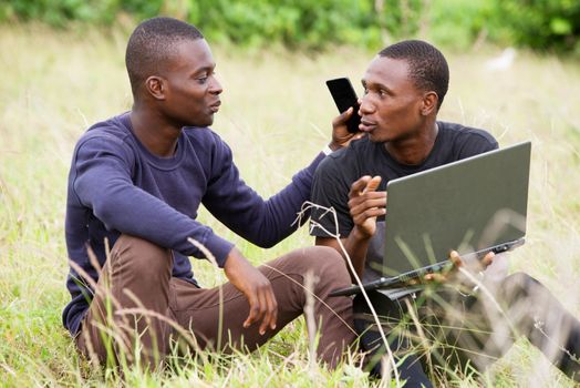two happy young men talk, work in front of a laptop and relax while sitting in the park.