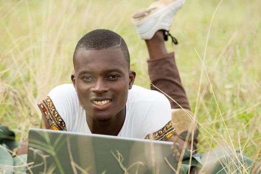 young man lying in grass in front of computer watching the camera smiling.