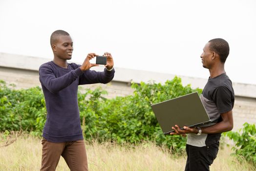 Young people standing in grass one with laptop in hand while the other makes him photo with mobile phone smiling.