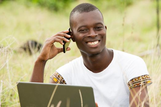 young man sitting in the grass in front of laptop and communicating with mobile phone laughing.