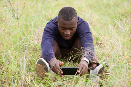 young smiling man sitting on the grass in the park is using and watching on mobile phone