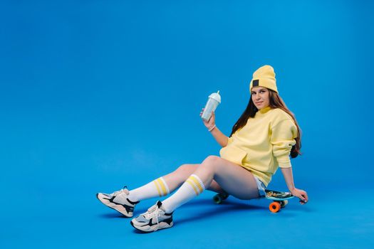 a pregnant girl in yellow clothes with a glass of juice sits on a skateboard on a blue background.