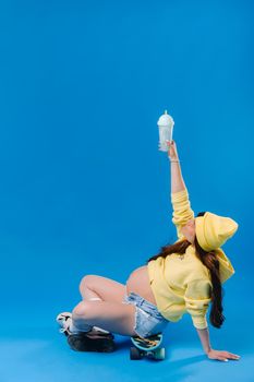 a pregnant girl in yellow clothes with a glass of juice sits on a skateboard on a blue background.