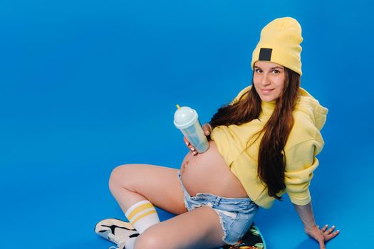 a pregnant girl in yellow clothes with a glass of juice sits on a skateboard on a blue background.
