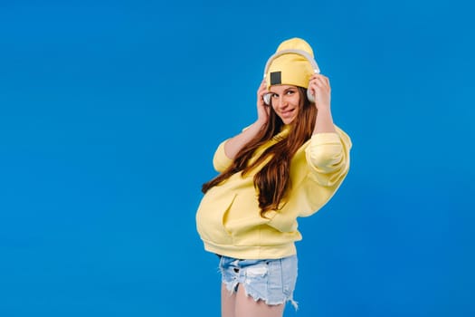 A pregnant girl in a yellow jacket and headphones stands on a blue background.