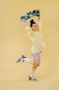 Pregnant girl in a yellow jacket with a skateboard in her hands on a yellow background.