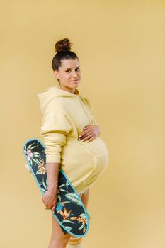 Pregnant girl in a yellow jacket with a skateboard in her hands on a yellow background.