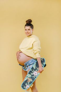 Pregnant girl in a yellow jacket with a skateboard in her hands on a yellow background.