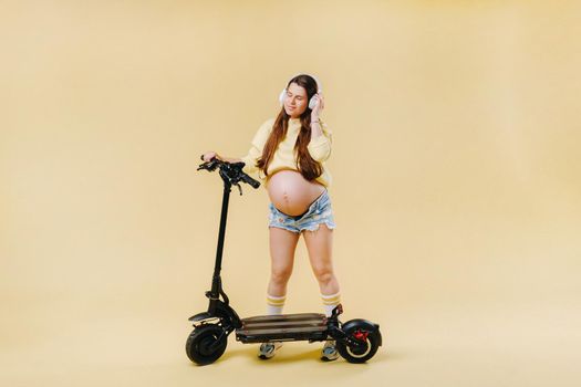 Pregnant girl in yellow clothes and headphones on an electric scooter on an isolated yellow background.