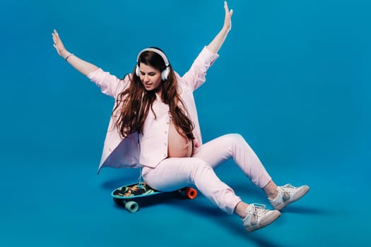 a pregnant woman in a pink suit sits on a skateboard with headphones and listens to music on a blue background.