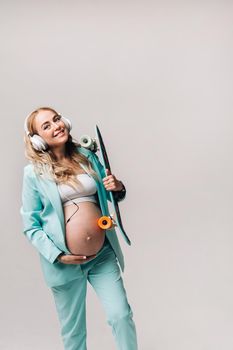 A pregnant girl in a turquoise suit with a skateboard in her hands and headphones stands on a gray background.