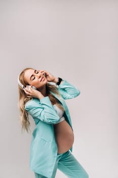 a pregnant woman in a turquoise suit with headphones stands and listens to music on a gray background.