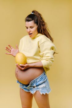 a pregnant girl with a yellow jacket stands with a melon in her hands on a yellow isolated background.