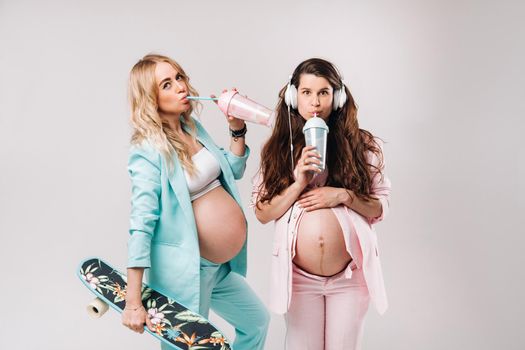 Two pregnant girls in turquoise and pink suits with glasses of juice, a skate and headphones stand on a gray background.