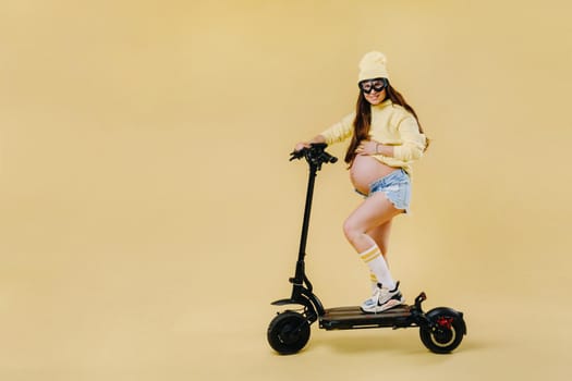 A pregnant girl in yellow clothes on an electric scooter on an isolated yellow background.