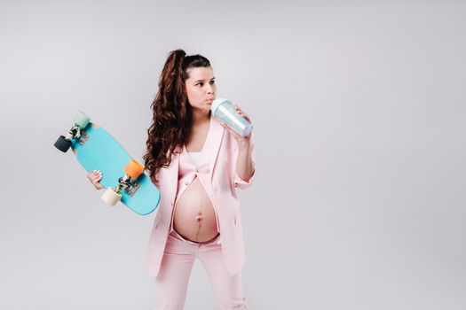 A pregnant girl in a pink suit with a skateboard in her hands drinks juice on a gray background.