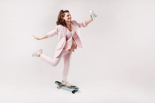 A pregnant girl in a pink suit with a skateboard in her hands stands on a gray background.