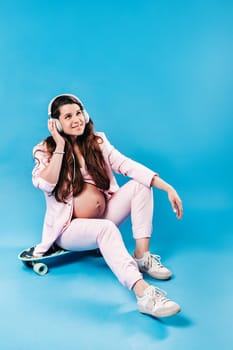 a pregnant woman in a pink suit sits on a skateboard with headphones and listens to music on a blue background.