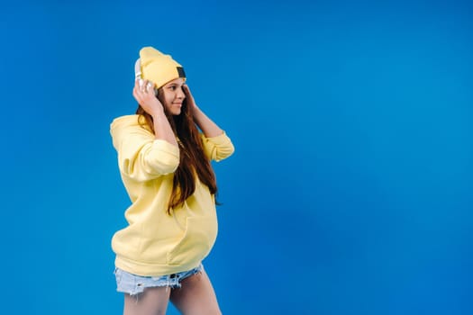 A pregnant girl in a yellow jacket and headphones stands on a blue background.