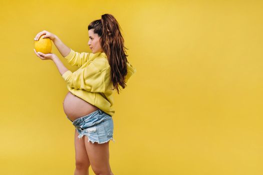 a pregnant girl with a yellow jacket stands with a melon in her hands on a yellow isolated background.